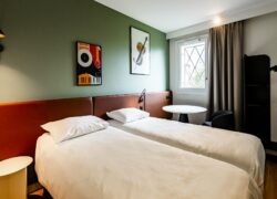 Une chambre d'hôtel avec deux lits simples, un mur vert et une tête de lit ocre, avec au mur deux tableaux sur représentant un piano et un violon.
