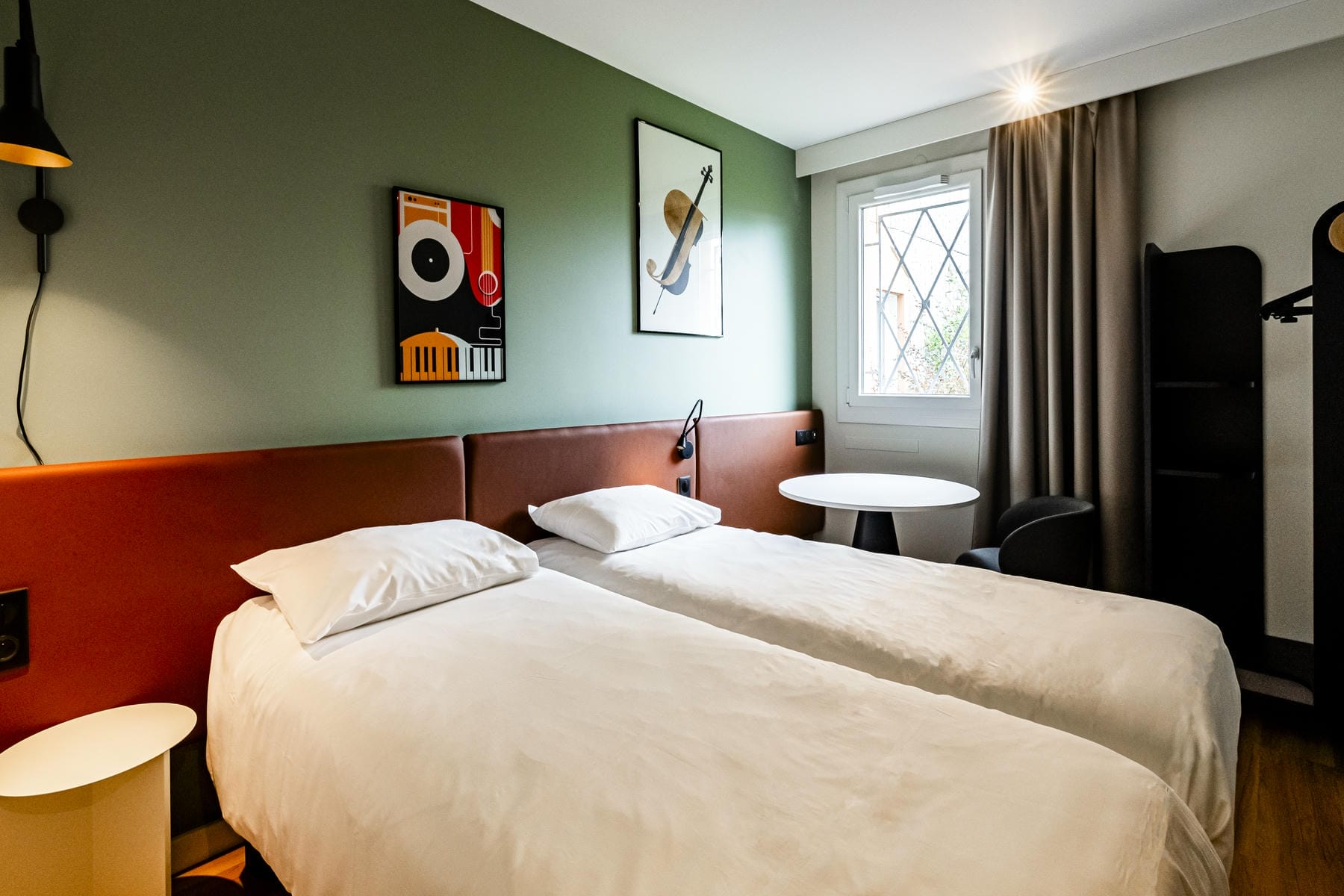 Une chambre d'hôtel avec deux lits simples, un mur vert et une tête de lit ocre, avec au mur deux tableaux sur représentant un piano et un violon.