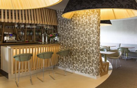 Bar aux tons doux de vert, beige et bois clair, avec comptoir rétroéclairé, tables dressées et cloison fleurie décorée d’un grand abat-jour suspendu au plafond en guise de luminaire.