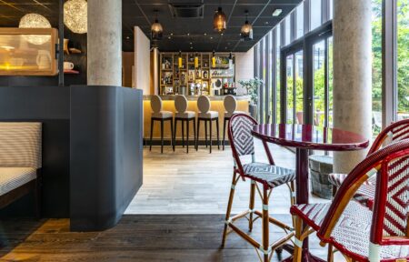 Un bar avec au premier plan un table bar et deux tabourets de bar rouge, et au fond, le bar et ses bouteilles
