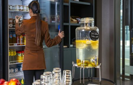 Un espace de pause avec devant une bonbonne d'eau infusée aux agrumes et des verres, et au fond une sihouette féminine qui ouvre un réfrigérateur vitré de la main droite et se sert d'un bocal gourmand de la main gauche