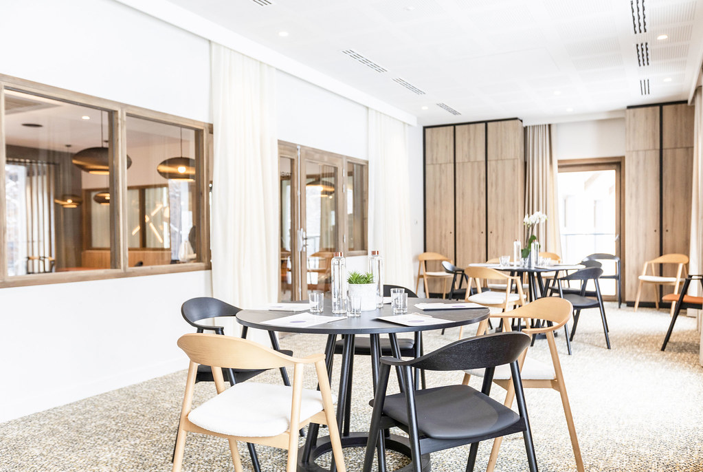 Salle de réunion moderne et lumineuse avec plusieurs tables rondes noires et des chaises mêlant bois clair et noir sur un sol clair moucheté, comprenant une table principale au premier plan dressée avec verres et bouteilles, d’autres tables disposées vers le fond, des salles vitrées éclairées chaleureusement visibles à gauche, et un long mur de panneaux et portes en bois clair à droite.