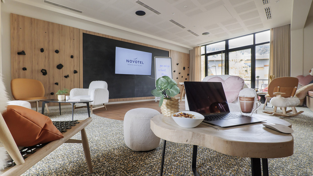 Salon et espace de réunion moderne d’hôtel avec des panneaux muraux en bois, un écran mural affichant le logo Novotel et des assises contemporaines disposées autour de tables basses, dont une table centrale avec un ordinateur portable, une boisson et une collation, à côté de grandes baies vitrées laissant entrer la lumière naturelle.