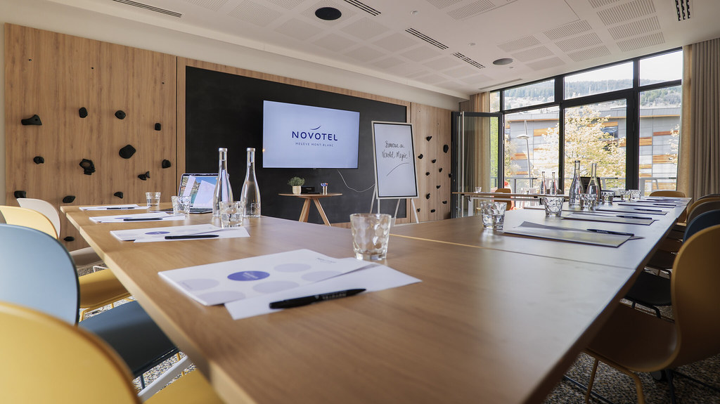 Salle de réunion d’hôtel moderne avec une longue table de conférence en bois dressée avec verres, bouteilles d’eau, carnets et stylos, faisant face à un écran mural affichant la marque Novotel, avec un paperboard et de grandes baies vitrées donnant sur des arbres et des bâtiments proches.