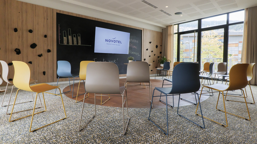 Salle de réunion moderne d’hôtel avec des chaises colorées disposées face à un espace de présentation comprenant un écran mural affichant “Novotel” et un mur type tableau noir, de grandes baies vitrées à droite laissant entrer la lumière naturelle et offrant une vue sur des arbres à l’extérieur.