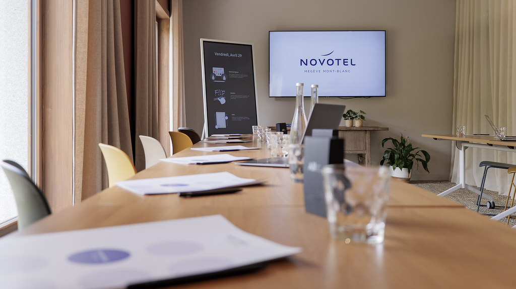 Salle de réunion d’hôtel avec une longue table en bois vue en perspective plongeante, disposée avec des documents, verres et bouteilles d’eau, bordée de chaises blanches et jaunes ; au fond, un écran mural affiche la marque Novotel à côté d’un panneau de présentation, tandis que des rideaux et une petite plante apportent une ambiance hôtelière moderne et chaleureuse.