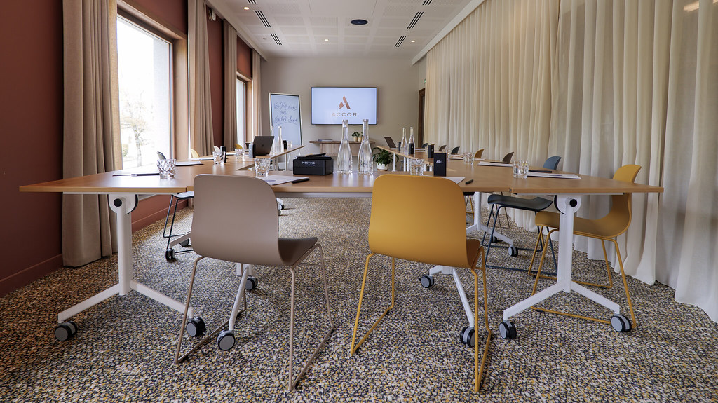 Salle de réunion moderne d’hôtel avec une grande table de conférence en bois entourée de chaises à roulettes de différentes couleurs, de verres et de matériel de présentation, un écran mural affichant le logo Accor et de hautes fenêtres avec rideaux sur un côté.
