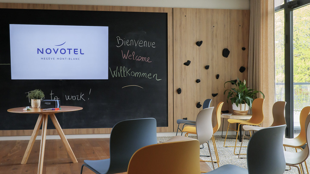 Salon d’hôtel moderne avec un grand mur en tableau noir affichant un écran “Novotel Megève Mont-Blanc” et les messages manuscrits “Bienvenue / Welcome / Willkommen”, comprenant des chaises colorées, une petite table ronde, des panneaux muraux en bois avec prises d’escalade et un coin assis avec des plantes près d’une fenêtre.