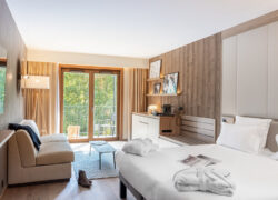 Chambre d’hôtel moderne avec mur d’accent en bois, comprenant à droite un lit double avec linge blanc, un espace bureau et coin café le long du mur du fond, un coin salon avec canapé et table basse à gauche, et de grandes portes vitrées ouvrant sur un balcon avec mobilier extérieur et vue sur la verdure.