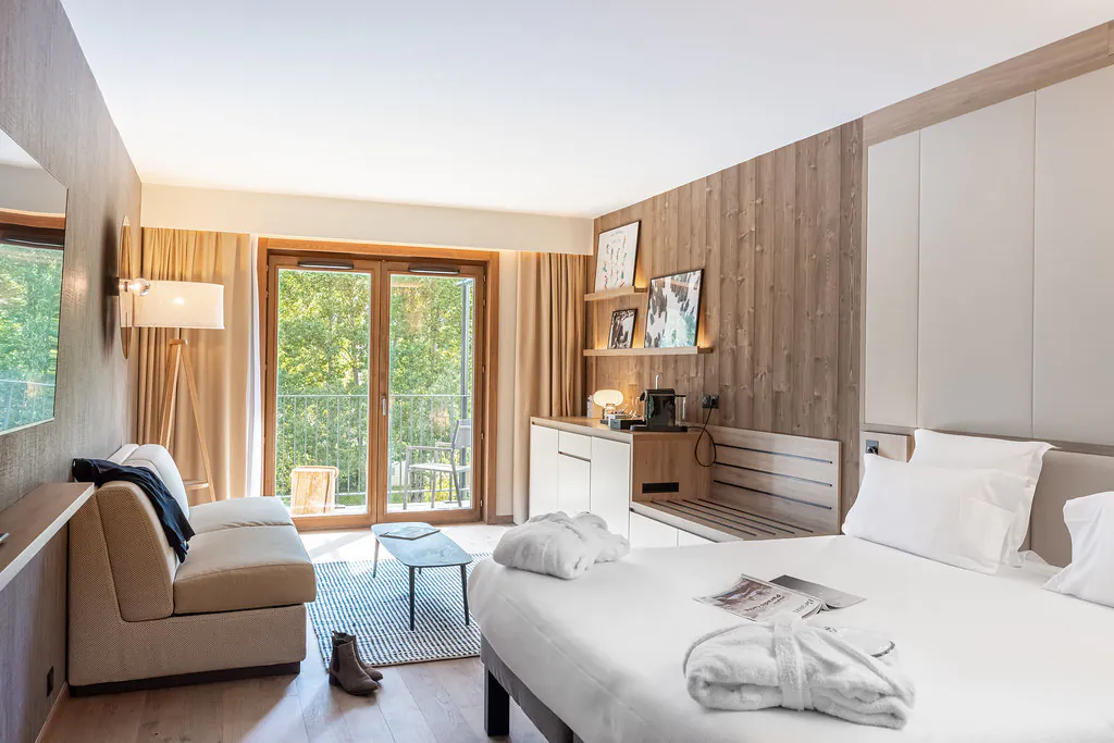 Chambre d’hôtel moderne avec mur d’accent en bois, comprenant à droite un lit double avec linge blanc, un espace bureau et coin café le long du mur du fond, un coin salon avec canapé et table basse à gauche, et de grandes portes vitrées ouvrant sur un balcon avec mobilier extérieur et vue sur la verdure.