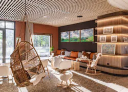 Salon d’hôtel moderne avec plafond en lattes de bois, fauteuil suspendu en osier au premier plan et coin détente composé d’un canapé orange, de fauteuils et d’une chaise à bascule près d’étagères intégrées éclairées et de tableaux de paysage, ouvert sur une grande baie vitrée donnant sur la verdure extérieure.