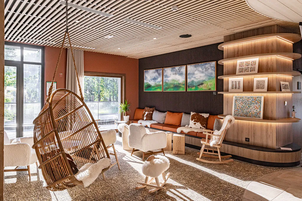 Salon d’hôtel moderne avec plafond en lattes de bois, fauteuil suspendu en osier au premier plan et coin détente composé d’un canapé orange, de fauteuils et d’une chaise à bascule près d’étagères intégrées éclairées et de tableaux de paysage, ouvert sur une grande baie vitrée donnant sur la verdure extérieure.