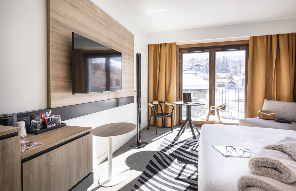 Chambre d’hôtel moquettée noir et blanc avec, à droite, un lit double faisant face à un écran de télévision, et au fond un coin salon composé d’une petite table ronde, de deux fauteuils et d’une banquette grise, devant une porte-fenêtre encadrée de rideaux orange ouvrant sur un balcon avec vue sur un village de montagne enneigé.