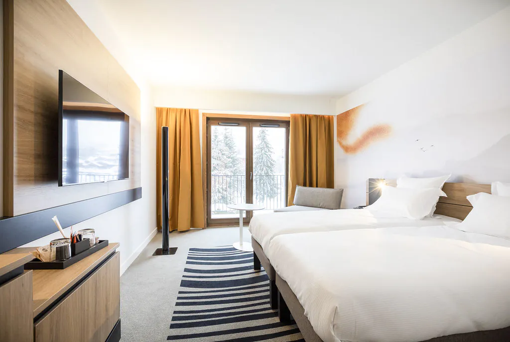 Chambre d’hôtel moquettée noir et blanc avec, à droite, deux lits twin faisant face à un écran de télévision, et au fond une porte-fenêtre encadrée de rideaux orange ouvrant sur un balcon avec vue sur des sapins enneigés.