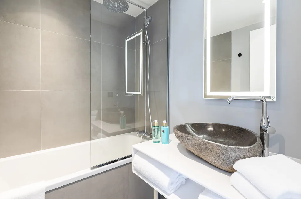 Gros plan sur un coin salle de bain avec, à gauche, une baignoire entourée d’un mur carrelé gris et d’une paroi vitrée, et à droite un lavabo en grès surmonté d’un miroir rectangulaire rétroéclairé, avec des serviettes blanches posées sur le lavabo et deux distributeurs de gel douche de la marque Rituals.