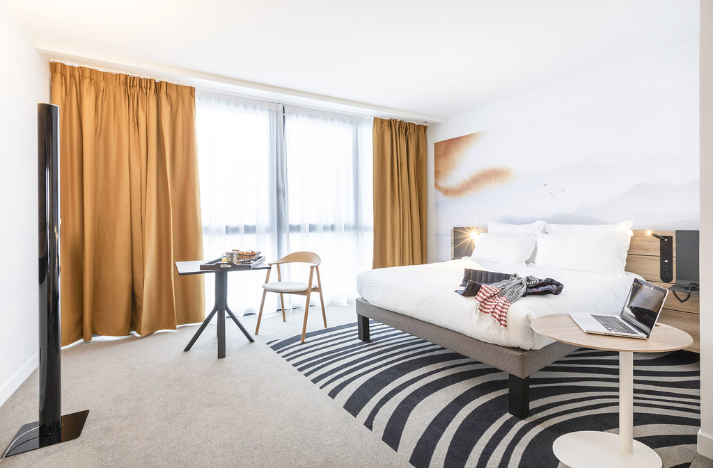 Chambre d’hôtel lumineuse et moquettée avec, à droite, un lit double et une petite table ronde sur laquelle est posé un ordinateur, et au fond à gauche une petite table carrée avec un plateau, un fauteuil, ainsi qu’une large baie vitrée encadrée de rideaux orange.