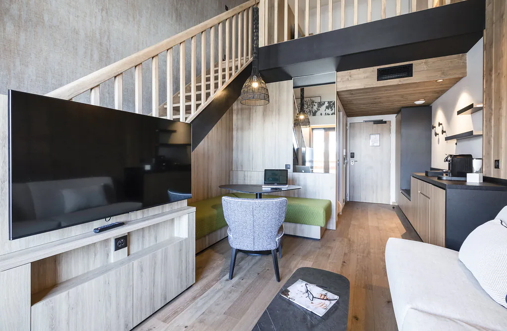 Salon aux murs en bois et parquet, décoré dans un style chalet moderne, avec au premier plan un coin salon comprenant une banquette et un écran TV, à gauche de la pièce un escalier menant à l’étage, au second plan un coin salle à manger, et au fond un couloir menant à l’entrée du duplex.