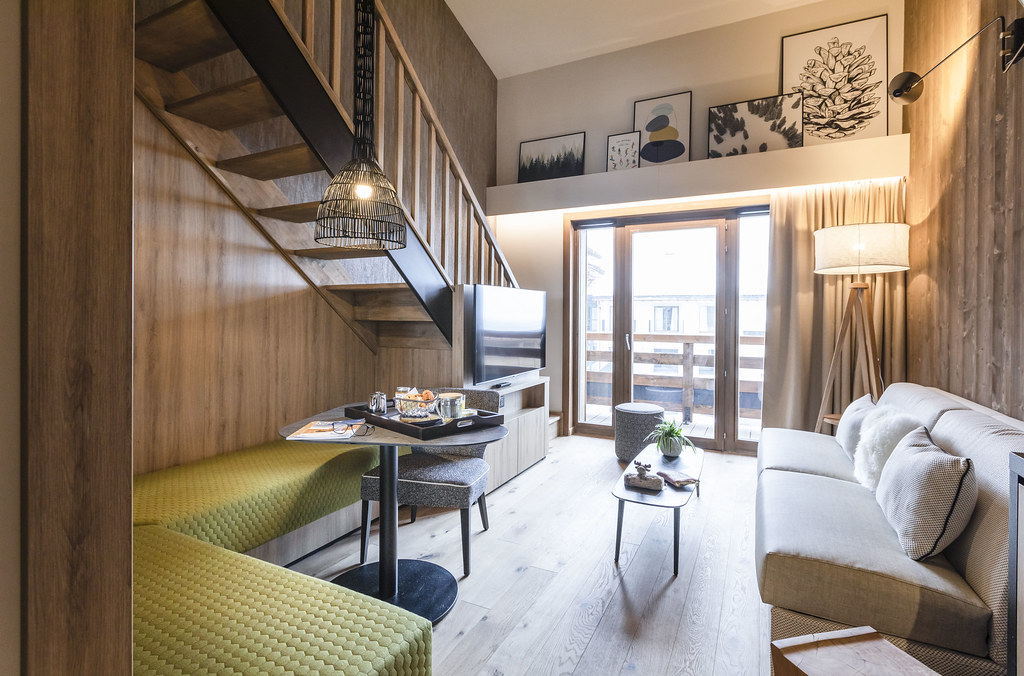 Salon aux murs en bois et parquet, décoré dans un style chalet moderne, avec un coin salle à manger à droite et un coin salon à gauche donnant sur un balcon en bois, et au fond de la pièce un escalier menant à l’étage.