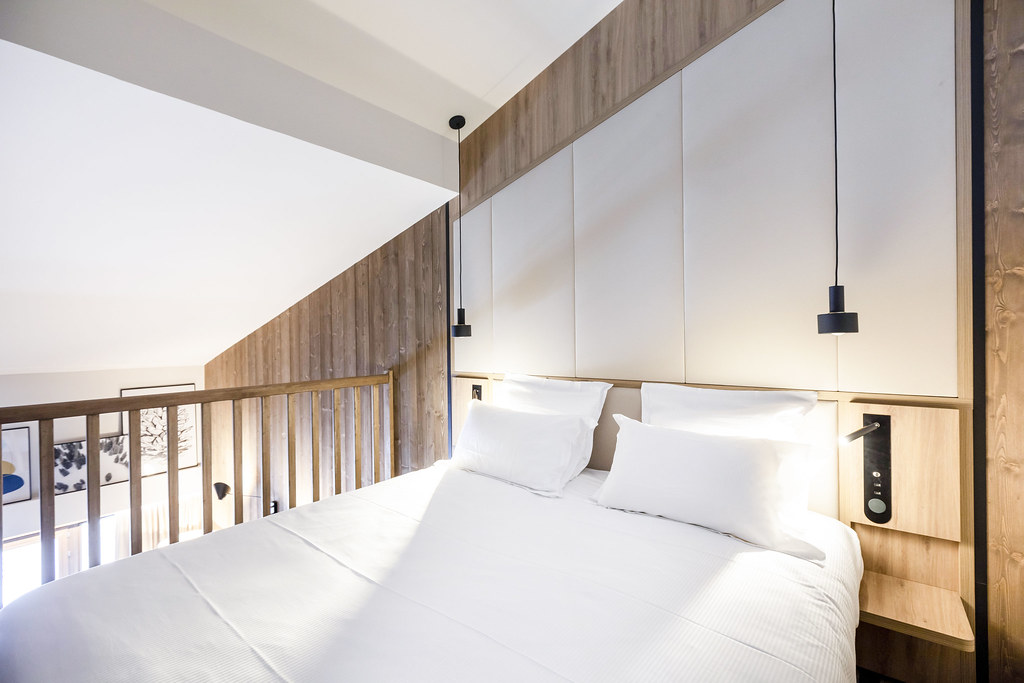 Chambre en mezzanine sous pente avec tête de lit capitonnée blanche, parement mural en bois et éléments de décoration modernes en aluminium noir.