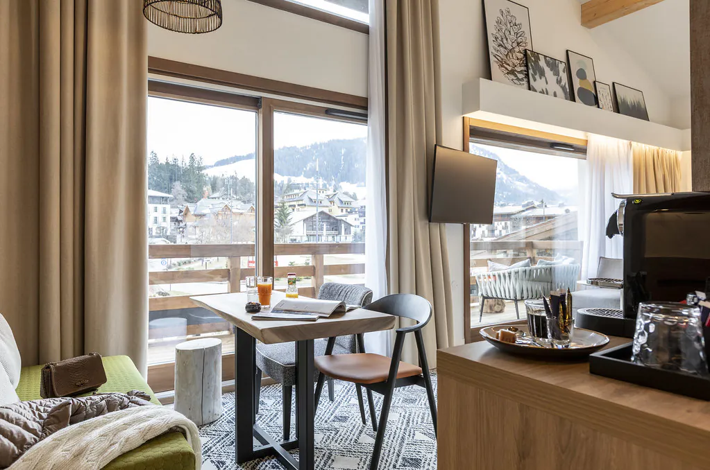 Intérieur aux hauts plafonds et moquetté, avec à gauche un coin salon comprenant une banquette verte, une table haute en bois et des fauteuils, à droite un meuble avec espace café, et sur toute la longueur une baie vitrée ouvrant sur un large balcon en bois aménagé offrant une vue sur le village et la montagne.