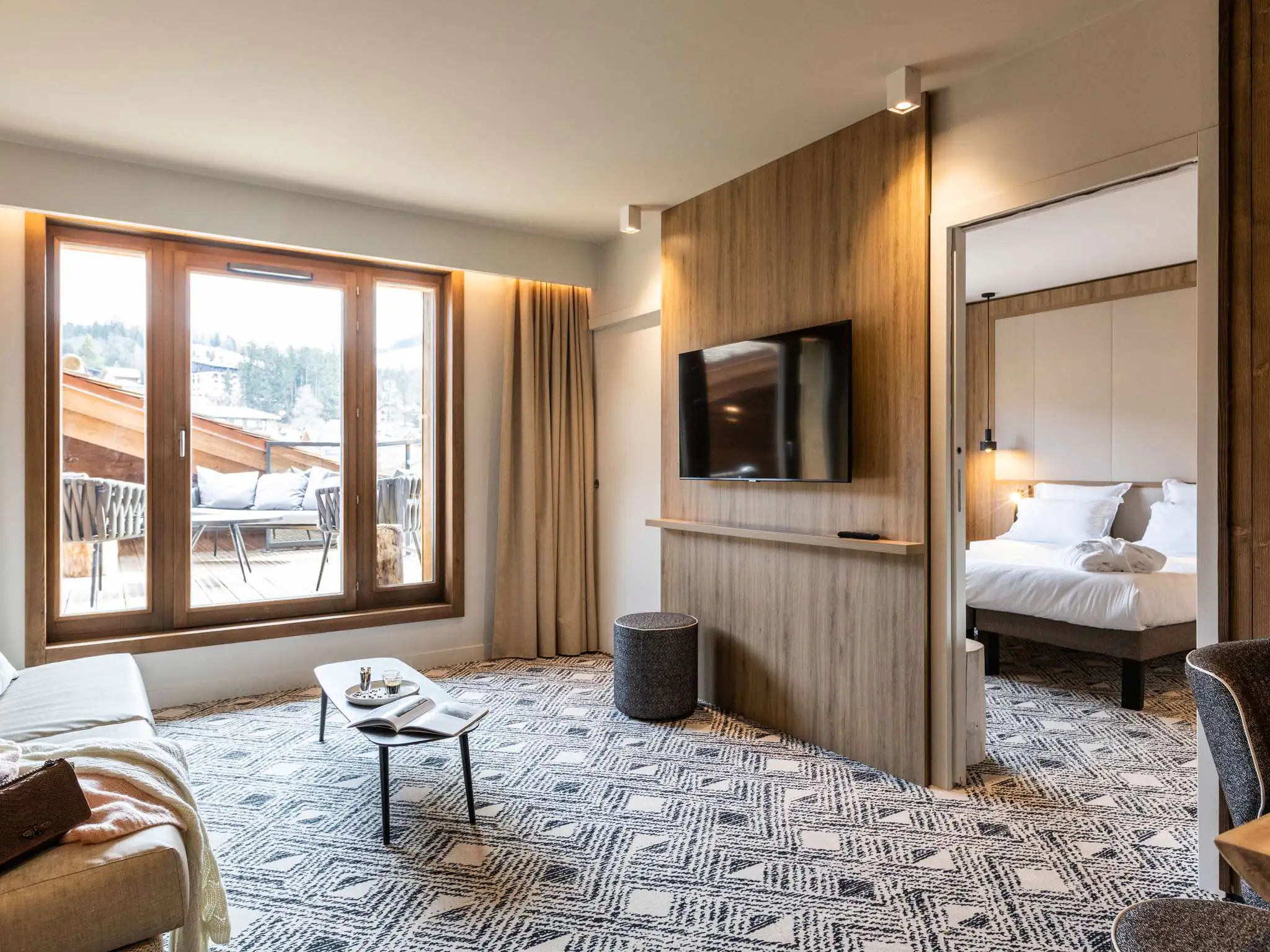 Chambre d’hôtel à la moquette noire et blanche et aux éléments muraux en bois, avec à gauche un coin salon composé d’une banquette et d’une table basse faisant face à un écran de télévision mural ; une baie vitrée ouvre sur une terrasse aménagée en salon de jardin avec vue sur les montagnes, tandis qu’à droite une ouverture mène à l’espace nuit avec un lit double et une haute tête de lit blanche capitonnée.