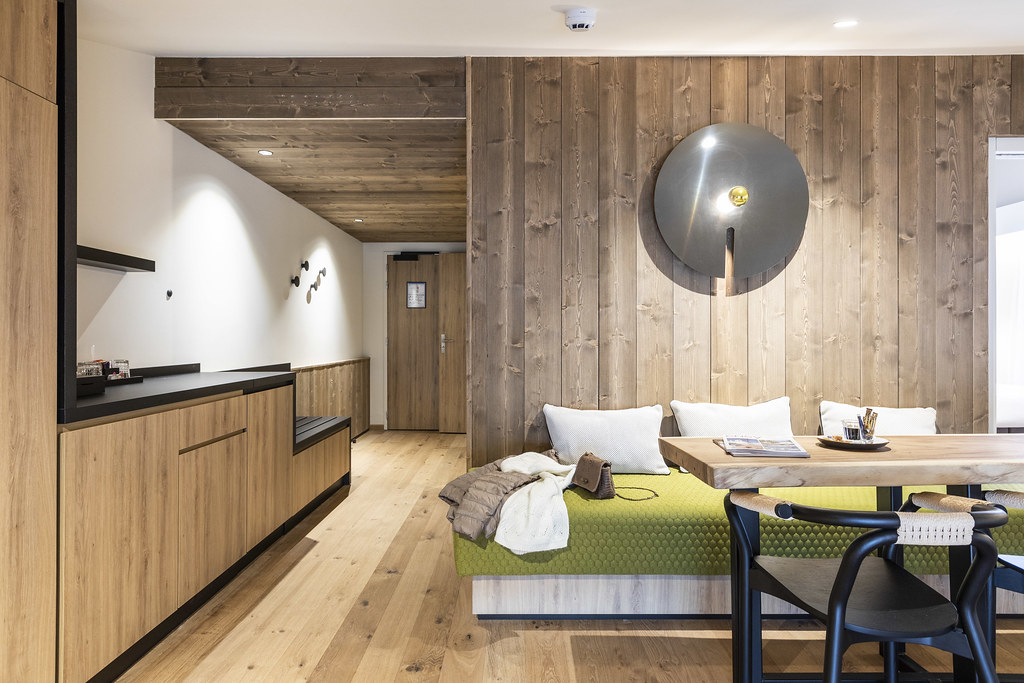 Coin salon d’une pièce habillée de bois avec, au fond à gauche, un couloir menant à l’entrée et à des espaces de rangement, et au premier plan à droite, un espace salle à manger composé d’une banquette verte, d’une table en bois brut et de fauteuils en métal noir et rotin.