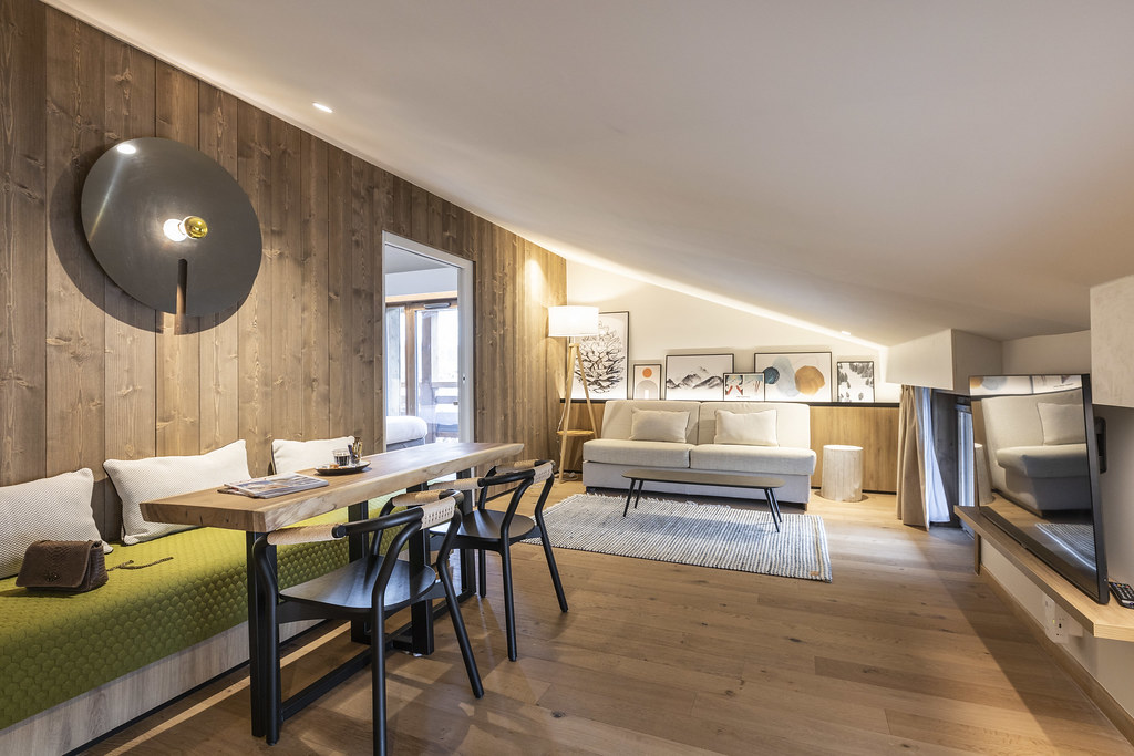 Salon mansardé habillé de bois au sol et sur les murs, avec à gauche un espace salon composé d’une table en bois brut, d’une banquette verte et de fauteuils en métal noir et rotin faisant face à un écran de télévision ; au fond de la pièce, une banquette blanche est décorée de plusieurs cadres muraux, à côté d’une ouverture laissant entrevoir la chambre adjacente.