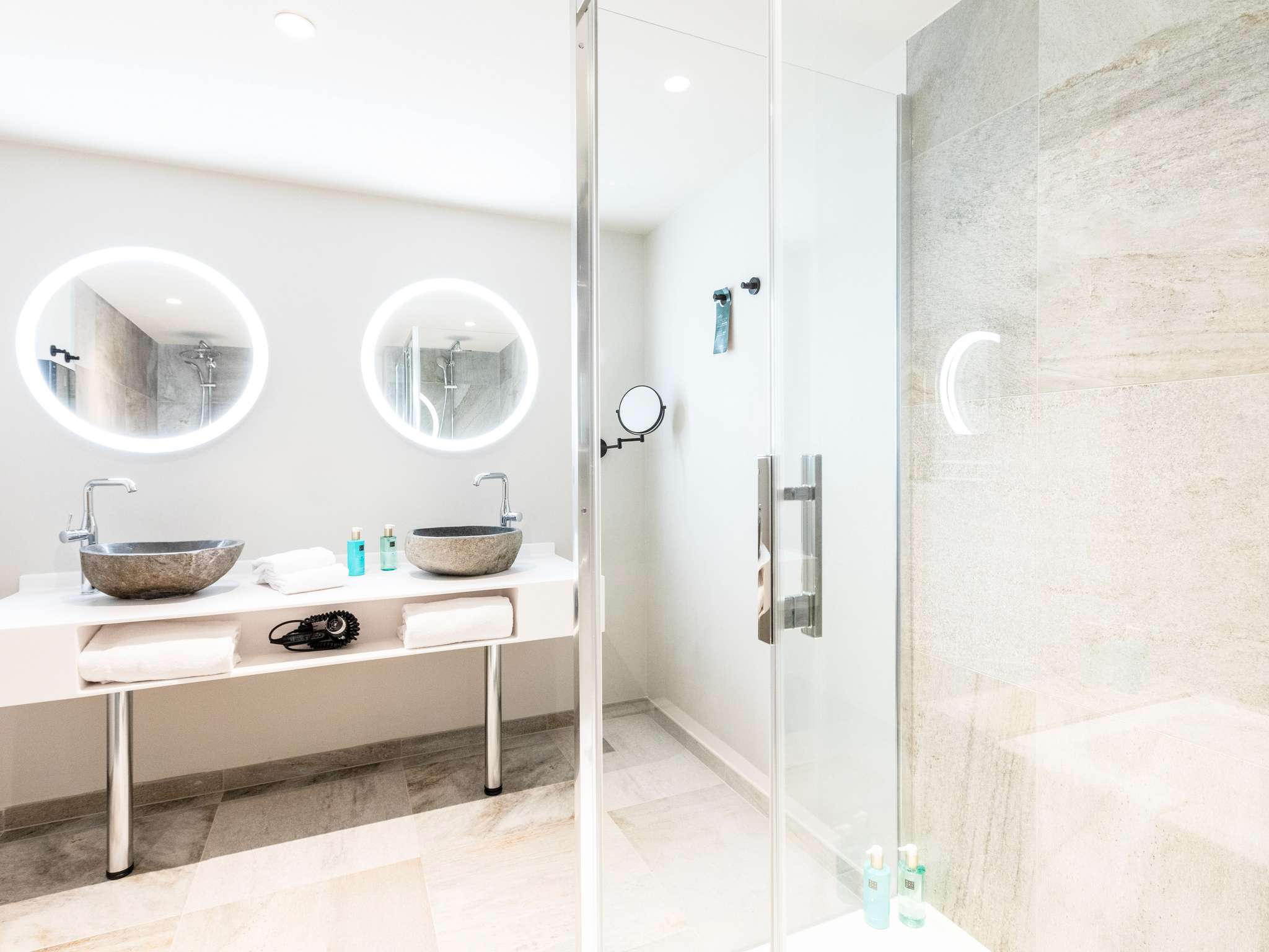 Salle de bain spacieuse et lumineuse avec, au premier plan, une paroi de douche vitrée et, au fond de la pièce, deux lavabos en grès surmontés de miroirs ronds rétroéclairés.