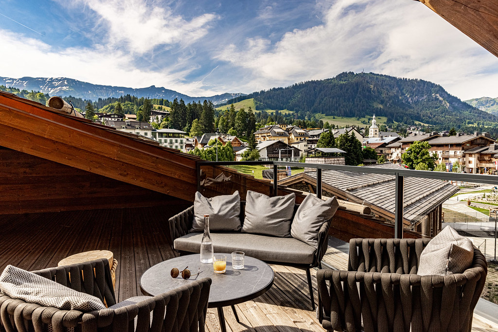 Terrasse en bois aménagée en salon de jardin avec vue dégagée sur les toits d’un village de montagne et, en arrière-plan, des sommets bordés de sapins.