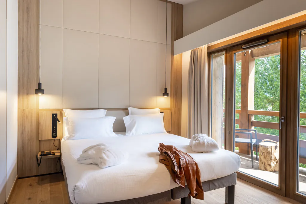 Chambre avec hauts plafonds et lit double central sur lequel sont posés deux peignoirs blancs et une veste en cuir marron, surmonté d’un mur en parement bois et d’une très haute tête de lit blanche capitonnée, et à droite une baie vitrée ouvrant sur un balcon en bois avec vue sur les arbres.