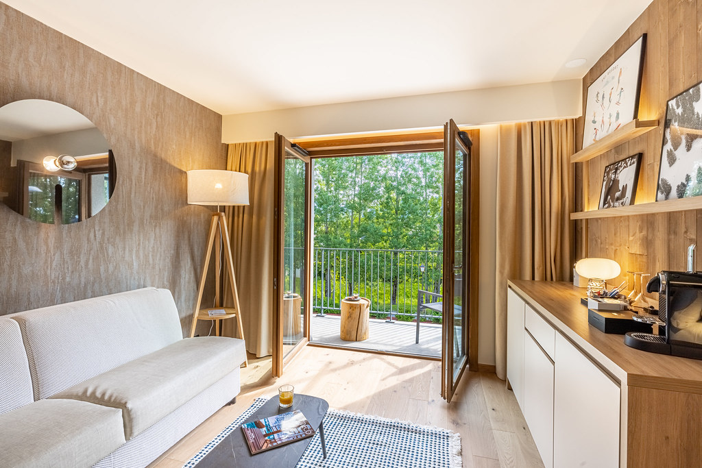 Coin salon habillé de bois avec, à gauche, une banquette blanche et une lampe sur pied en bois, une table basse, et à droite un meuble en bois avec divers éléments de décoration et une machine à café ; au fond, une grande porte vitrée ouverte sur un balcon offrant une vue sur les arbres.