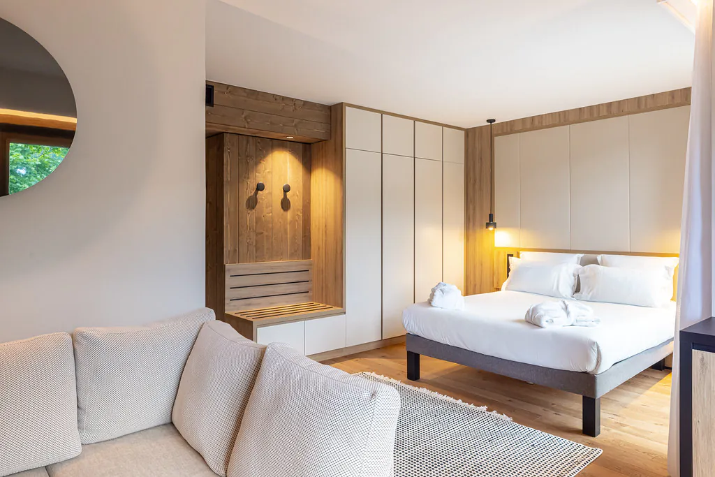 Chambre d’hôtel avec, à gauche, un bout de banquette crème surmontée d’un miroir, et à droite un lit avec deux peignoirs posés dessus et un tapis au pied sur parquet ; le lit est surmonté d’une tête de lit capitonnée, et au fond de la pièce se trouvent des rangements toute hauteur en bois et blanc.