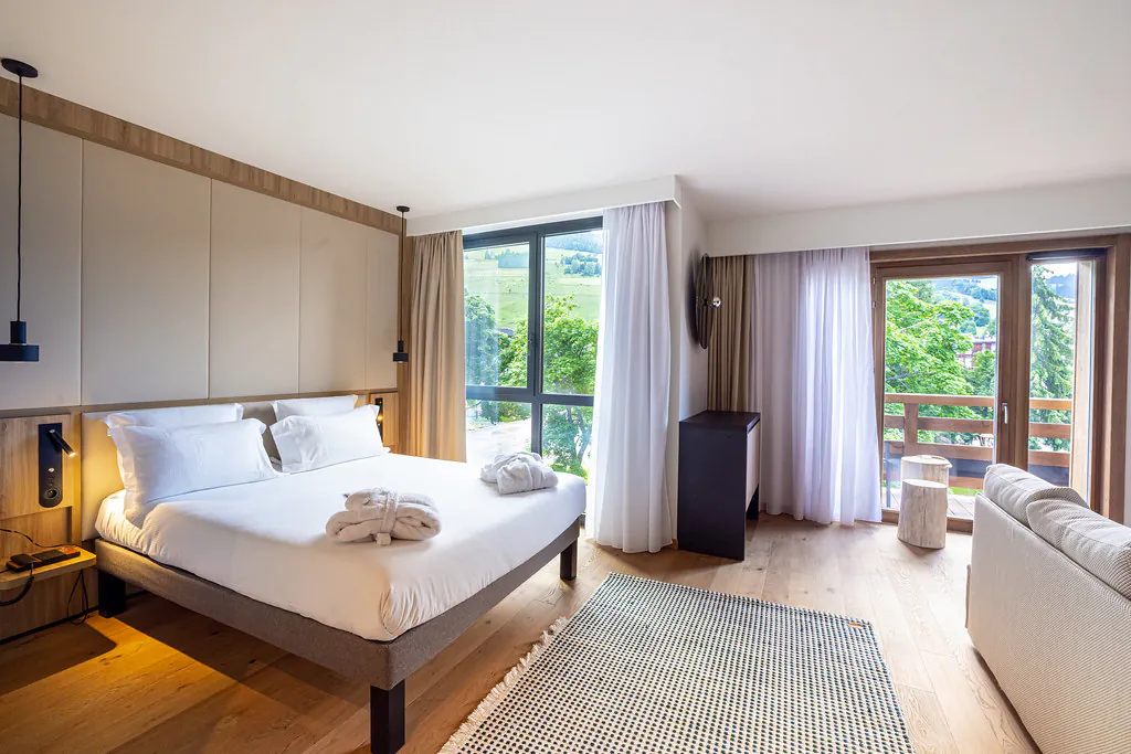 Grande chambre avec, à droite, un lit double sur lequel sont posés des peignoirs et surmonté d’une tête de lit capitonnée ; à son pied, un grand tapis sur parquet, et à droite une banquette, l’ensemble étant ouvert sur des fenêtres donnant sur un balcon en bois avec vue sur les arbres.