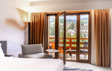 Chambre avec un lit et un fauteuil gris, donnant sur une porte-fenêtre ouvrant sur un balcon aménagé avec vue sur un village de montagne.