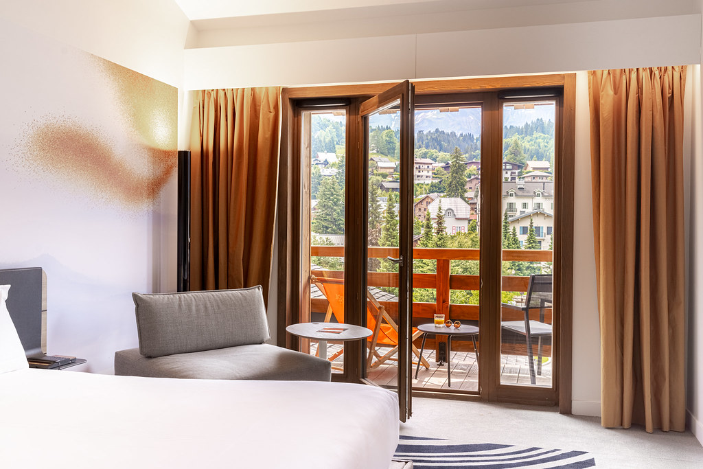 Chambre avec un lit et un fauteuil gris, donnant sur une porte-fenêtre ouvrant sur un balcon aménagé avec vue sur un village de montagne.