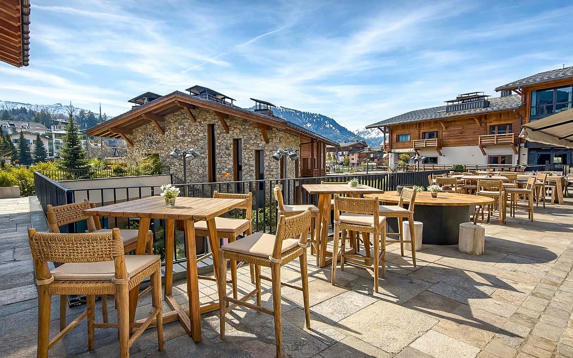 Grande terrasse de restaurant dallée avec garde-corps en aluminium noir et mobilier en bois, offrant une vue sur des chalets et un village de montagne, avec des sapins et des sommets enneigés en arrière-plan.