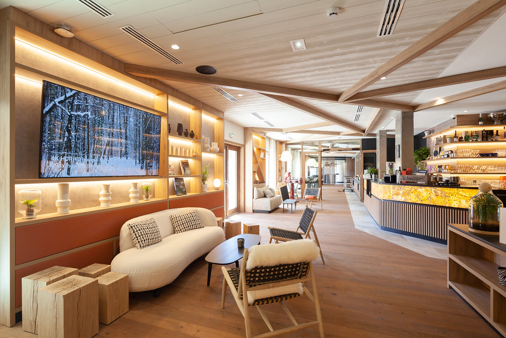 Grand espace salon commun coloré et lumineux à dominante de bois clair, avec au premier plan plusieurs assises confortables disposées autour d’une table basse sous un grand écran de télévision, à droite au second plan un bar rétroéclairé, et sur toute la longueur de la pièce diverses assises jusqu’au fond de la salle.