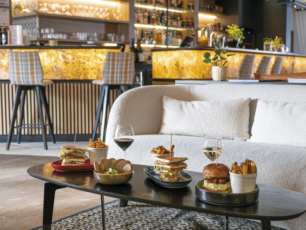 Gros plan sur une table basse en bois noir devant une banquette en bouclette blanche, sur laquelle sont posées plusieurs assiettes de finger food (club sandwichs, burgers, frites, tartinables et tranches de pain) accompagnées de deux verres de vin, avec en arrière-plan un bar et plusieurs tabourets hauts à motif écossais.
