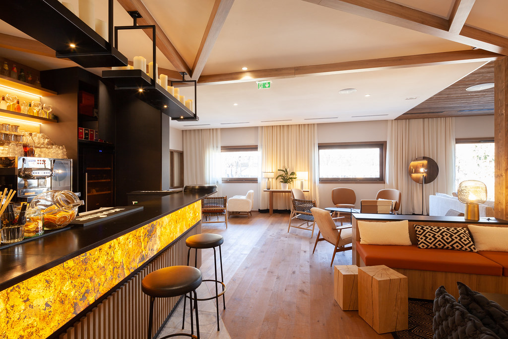 Espace lounge habillé de bois avec, à droite, un bar en bois sombre rétroéclairé, un espace avec banquette et assises en bois brut, et au fond des salons composés de fauteuils autour de tables basses, l’ensemble ouvert sur de larges fenêtres encadrées de rideaux blancs.