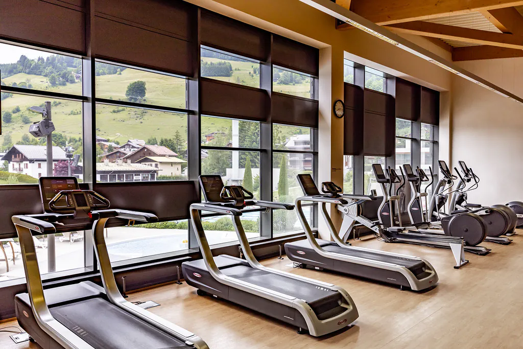 Espace cardio d’une salle de sport moderne avec plusieurs tapis de course et vélos elliptiques alignés devant de grandes baies vitrées offrant une vue sur des collines verdoyantes et des habitations en montagne.