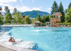 Piscine extérieure dans une station de montagne avec une eau bleue claire, une femme se détendant au bord de l’eau au premier plan, et une tour de surveillance en bois, des espaces de détente et des montagnes boisées à l’arrière-plan sous un ciel ensoleillé.