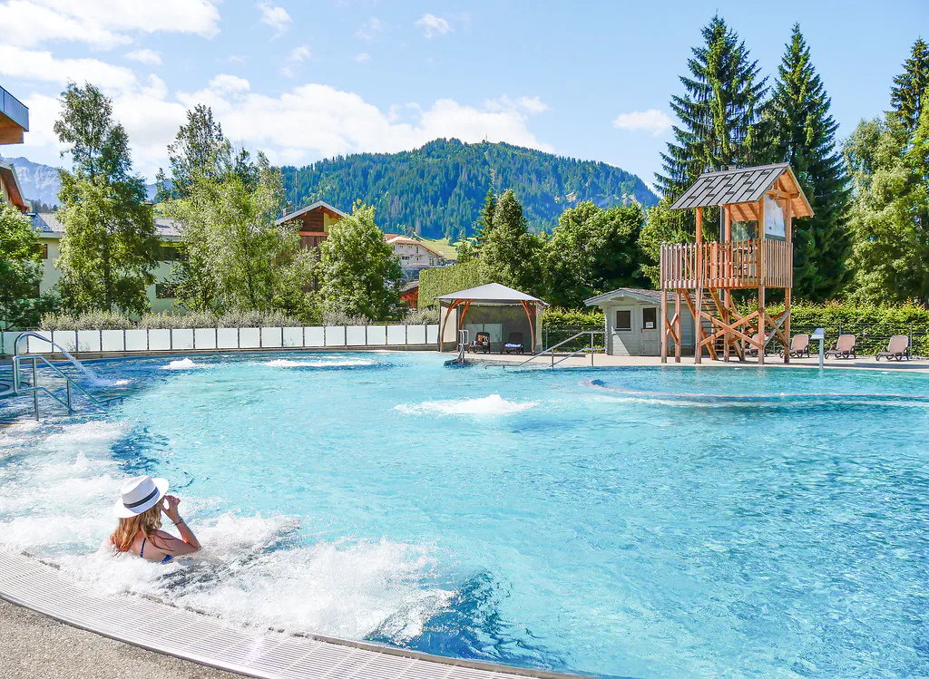 Piscine extérieure dans une station de montagne avec une eau bleue claire, une femme se détendant au bord de l’eau au premier plan, et une tour de surveillance en bois, des espaces de détente et des montagnes boisées à l’arrière-plan sous un ciel ensoleillé.