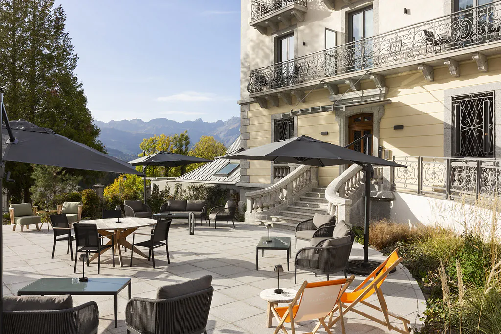 Exterior view of a historic building with, in the foreground, a terrace arranged as an outdoor lounge featuring tables, armchairs, and parasols, and in the background, a mountain landscape.