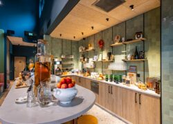 Buffet petit déjeuner en arrondi, mur carrelé vert amande, plafond en liège et grande table au premier plan avec fruits, pains et bonbonne de thé.