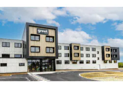 Au premier plan, une route qui dessert un hôtel dont la façade cubique et moderne de couleurs noire, blanche et bois clair s'élève au second plan.