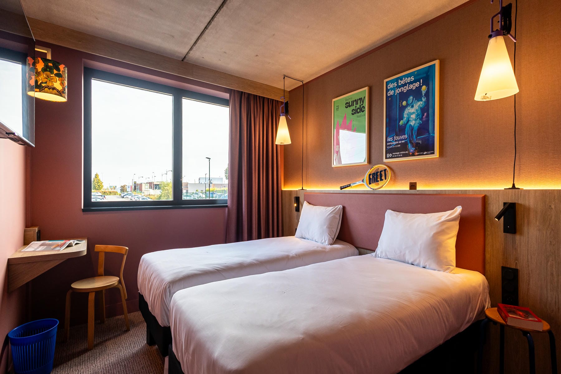 Chambre avec lits twin, murs orangés et vieux rose, luminaires jaunes et poster représentant une personne qui jongle, dans une atmosphère très peps.