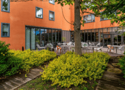 Façade orange de l’hôtel avec baies vitrées en angle au rez-de-chaussée, terrasse en bois dressée et végétation environnante.