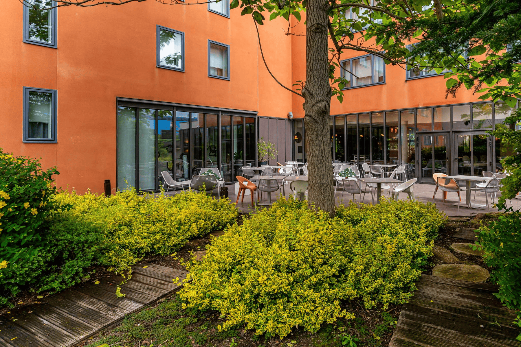 Orange hotel façade with corner bay windows on the ground floor, a set wooden terrace, and surrounding greenery.