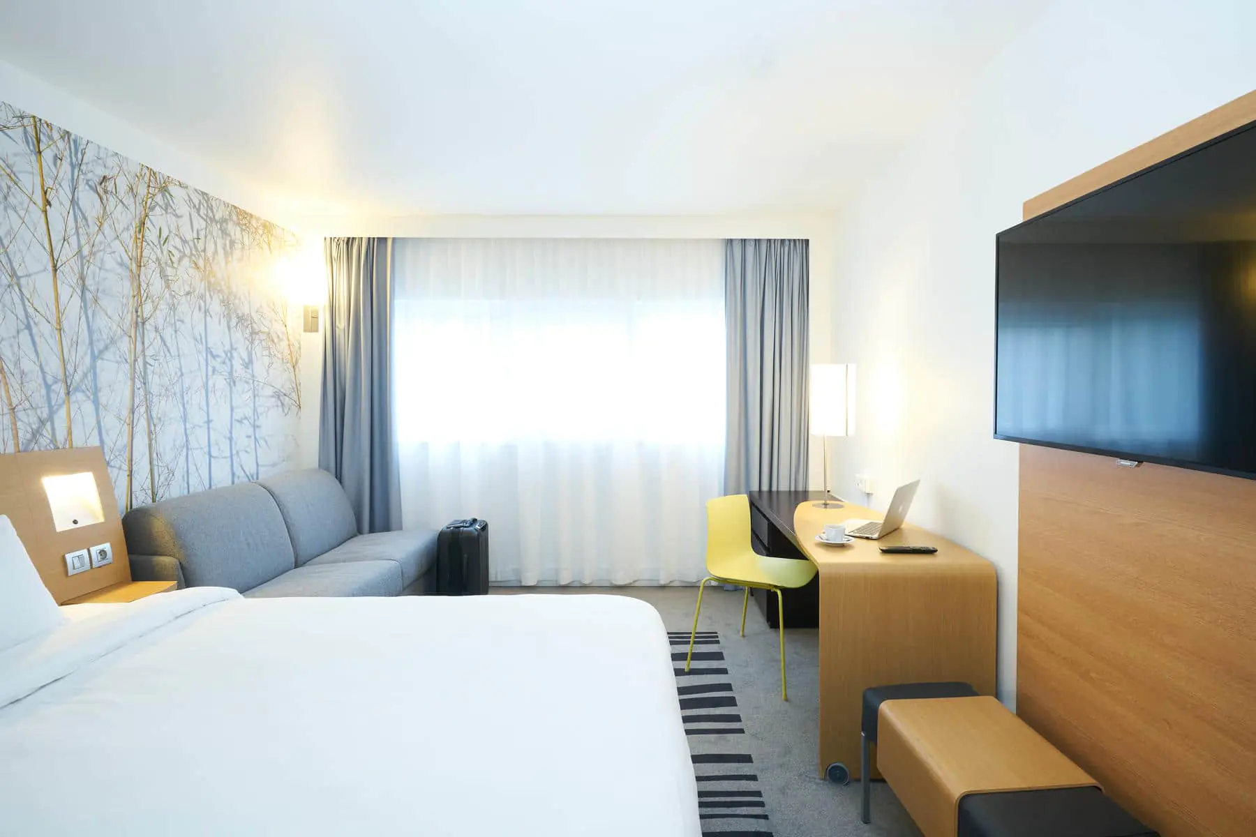 A bright grey-toned bedroom with a double bed on the left facing a wall-mounted flat-screen TV, and at the back of the room a grey sofa on the left, a workspace on the right, and a large window in the background.