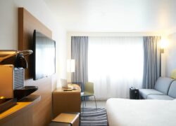 Une chambre lumineuse avec à droite un grand lit double qui fait face à une machine à café et à une télé écran plat au mur et au fond de la pièce un canapé gris sur la droite et un coin bureau sur la gauche, et une large fenêtre à l'arrière plan.